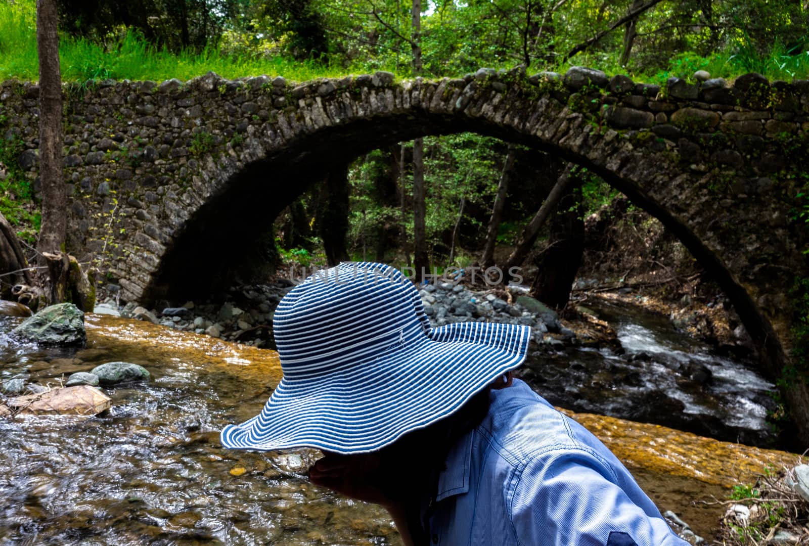 A girl in a wide-brimmed hat drinks water from a stream next to a medieval Venetian bridge in the center of the island of Cyprus.