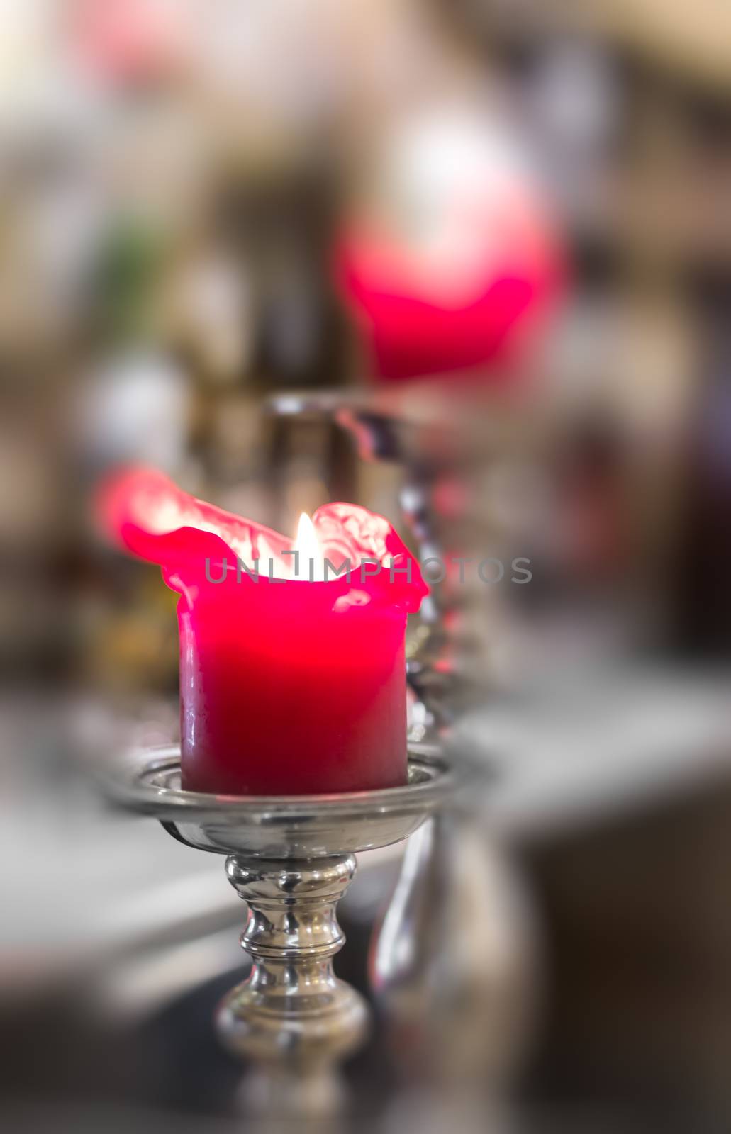 Close-up detail of red candle in silver candlestick lighting up a church. On Defocused blurry background they can be seen other red candle. Symbol of religion, prayer and peace.