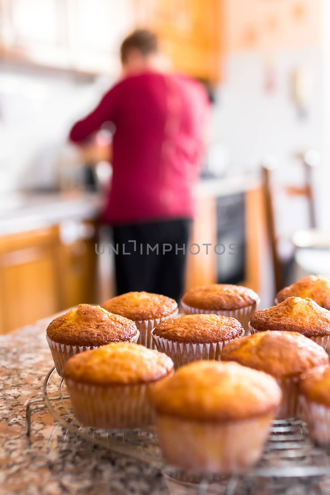 Batch of homemade freshly baked cupcakes or muffins cooling on a wire rack in the kitchen in a close up view with selective focus. Defocused blurry background. In the background the grandmother who has just cooked them.