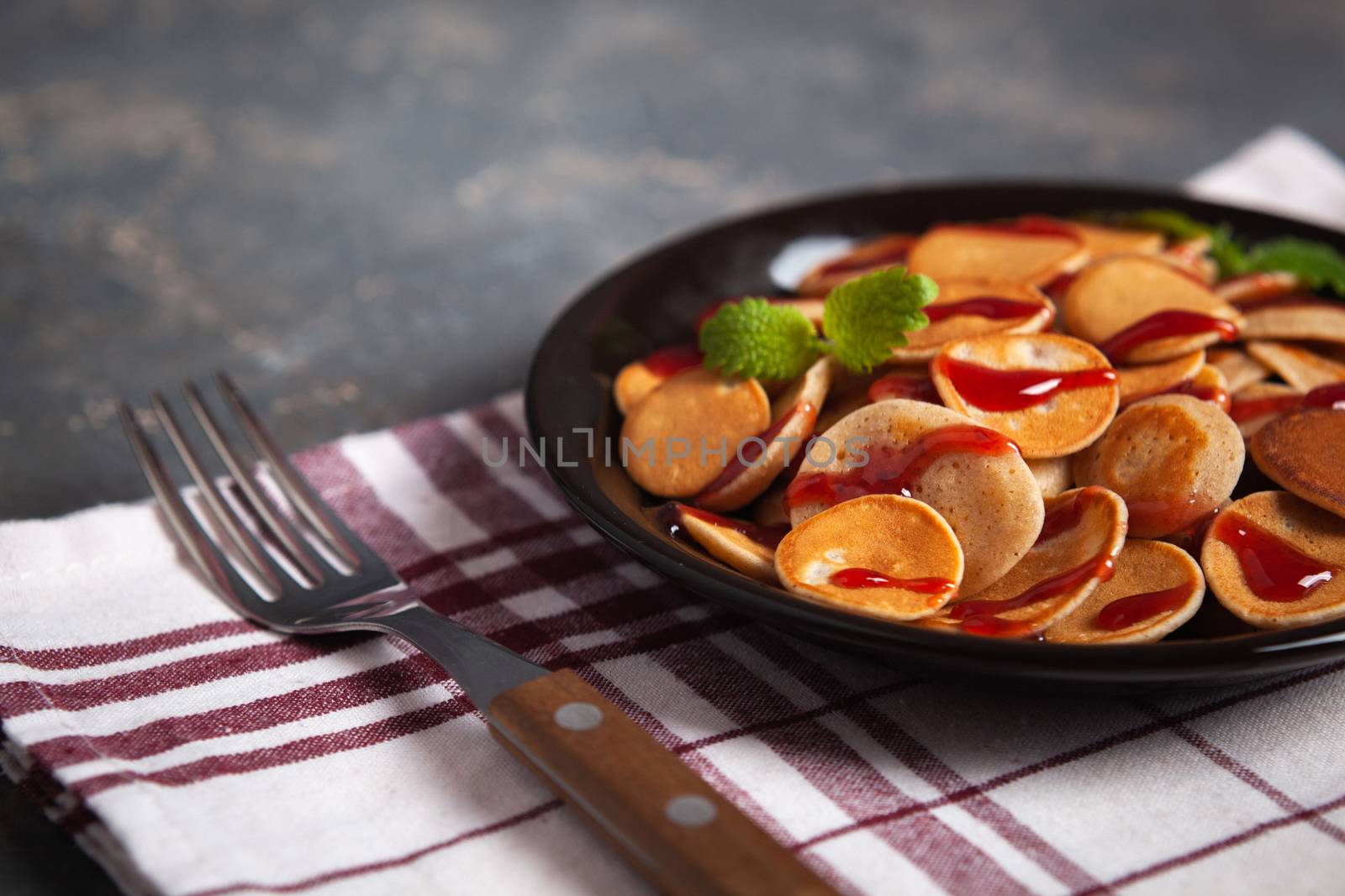 Trendy homemade breakfast, pancake cereal, mini pancakes on dark plate on grunge gray concrete background with check towel and fork, leaves of lemon balm or peppermint and cherry jam. Space for text.