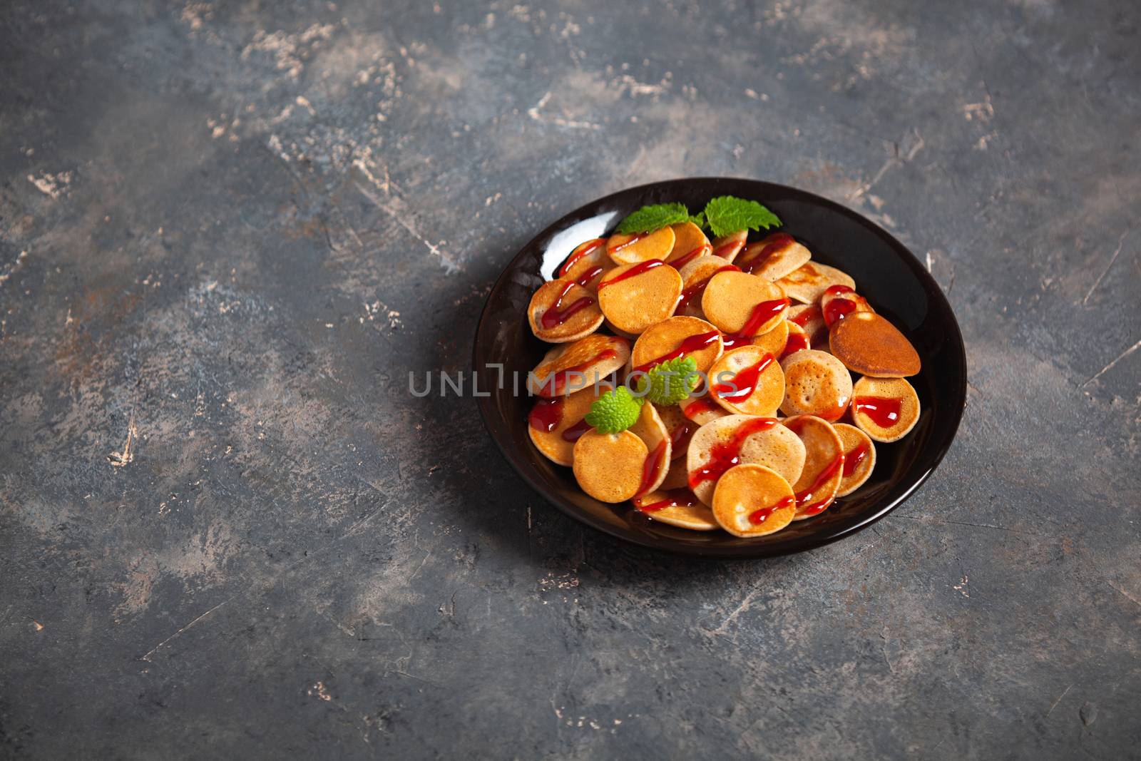 Trendy homemade breakfast, pancake cereal, mini pancakes on dark plate on grunge gray concrete background, leaves of lemon balm or peppermint and cherry jam. Space for text.