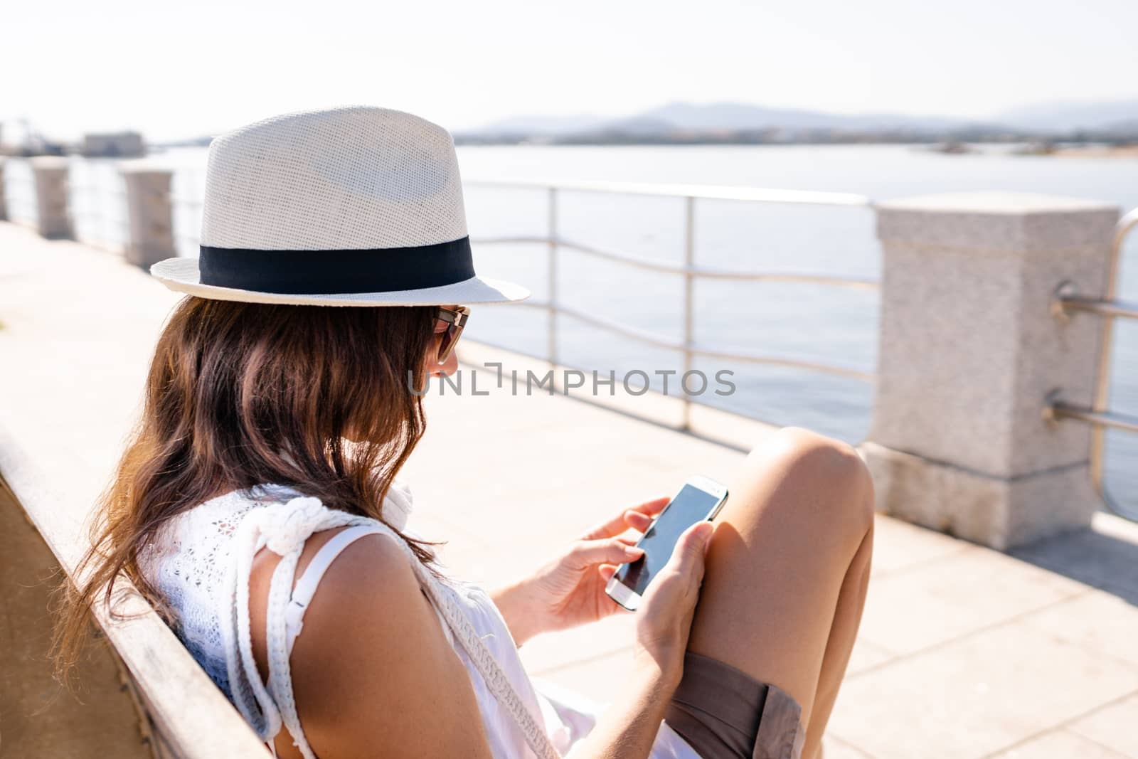 Caucasian young woman with white hat sitting on a bench of a sidewalk by the sea using her smartphone - Single woman outdoor connected with new social technology by robbyfontanesi