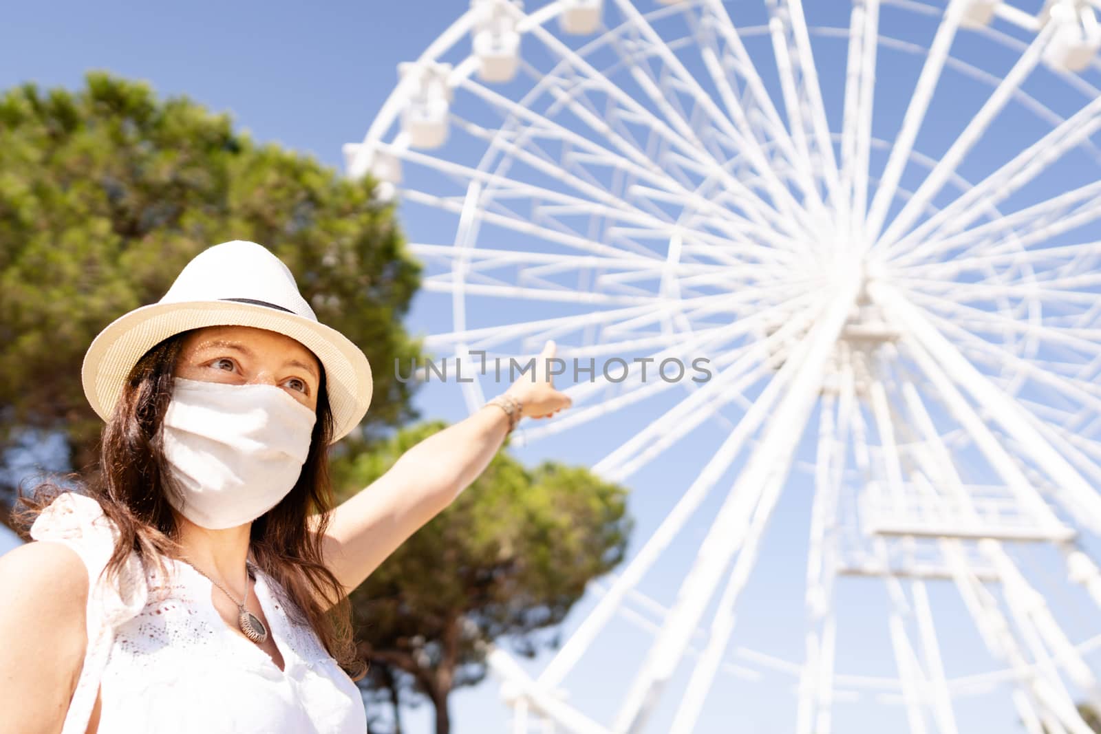 Caucasian young woman with white hat and COVID mask protection pointing to the ferris wheel - New tourism rules - Summer vacation with Coronavirus safe concept - Female tourist with protection mask by robbyfontanesi
