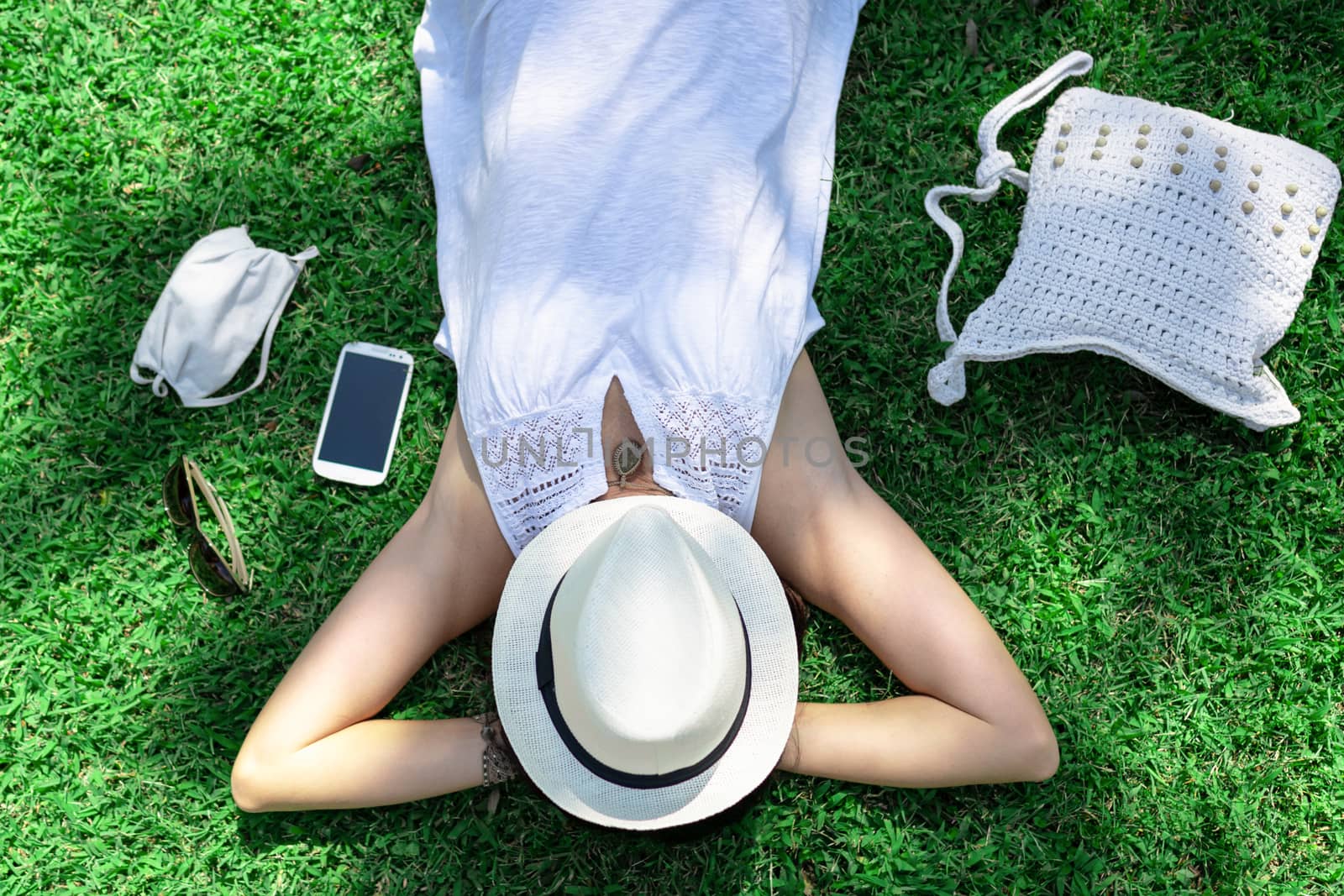 Top view of a Caucasian woman resting lying on a green field of the park with the face covered by her white hat her white Panama hat and COVID protection mask next to it - City vacation for safety by robbyfontanesi