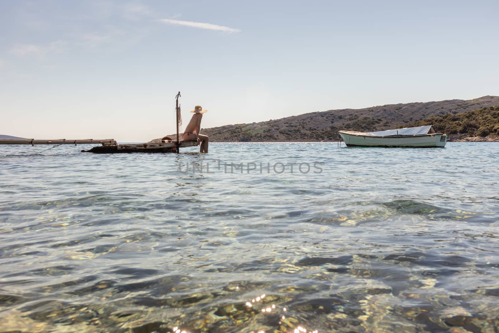 View of unrecognizable woman wearing big summer sun hat tanning topless and relaxing on old wooden pier in remote calm cove of Adriatic sea, Croatia by kasto