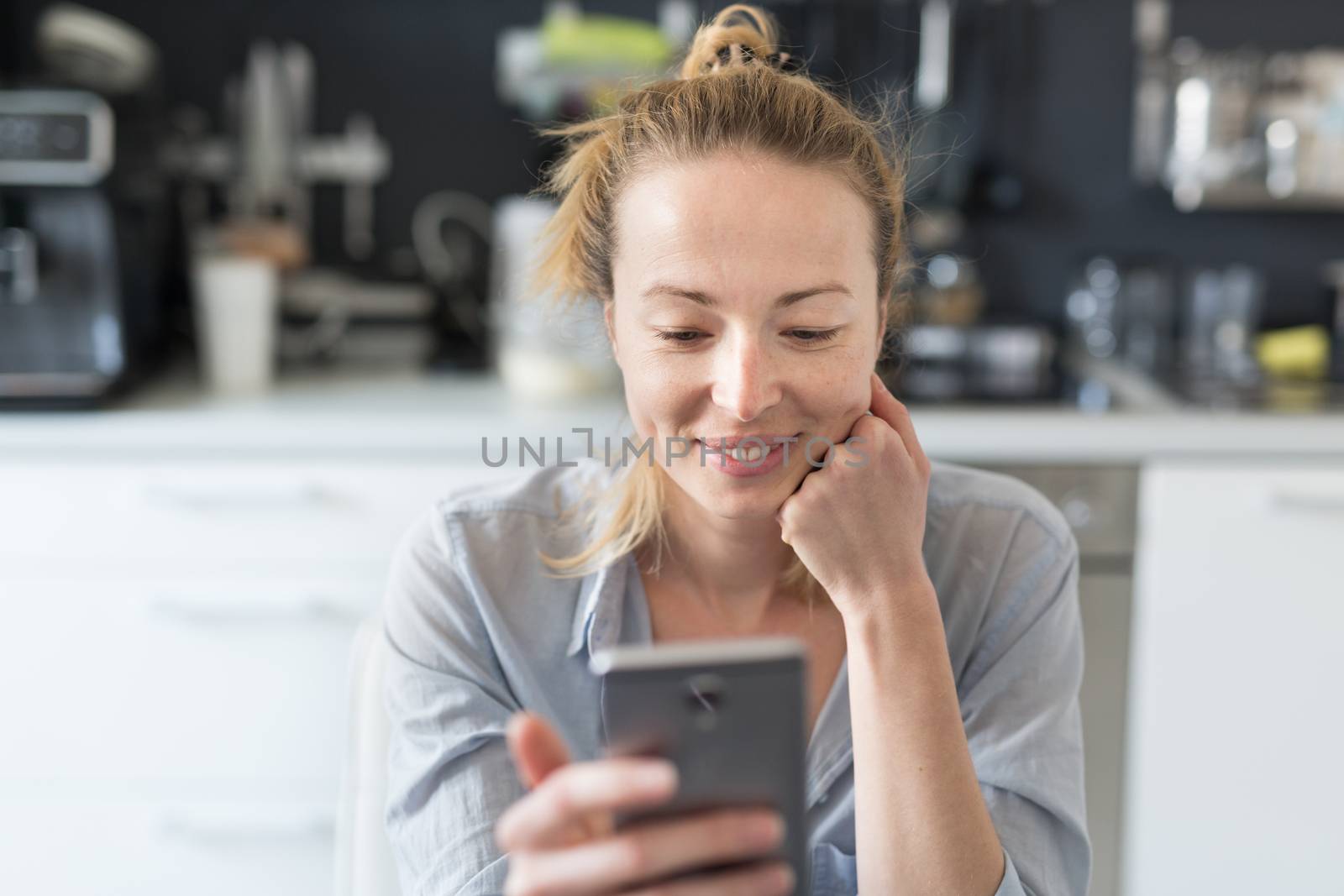 Young smiling cheerful woman indoors at home kitchen using social media apps on phone for video chatting and stying connected with her loved ones. Stay at home, social distancing lifestyle.
