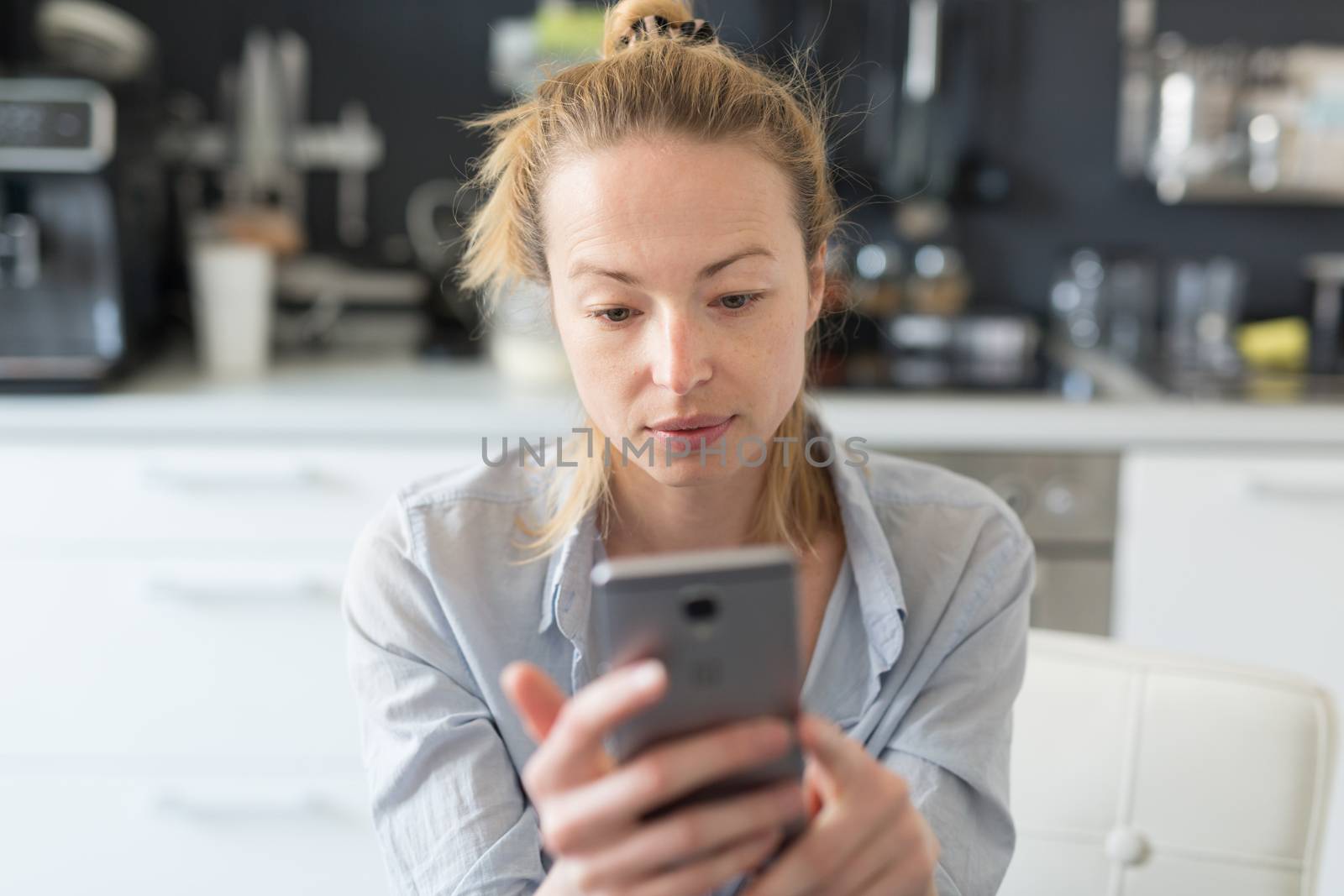 Worried woman indoors at home kitchen using social media apps on phone for video chatting and stying connected with her loved ones. Stay at home, social distancing lifestyle.