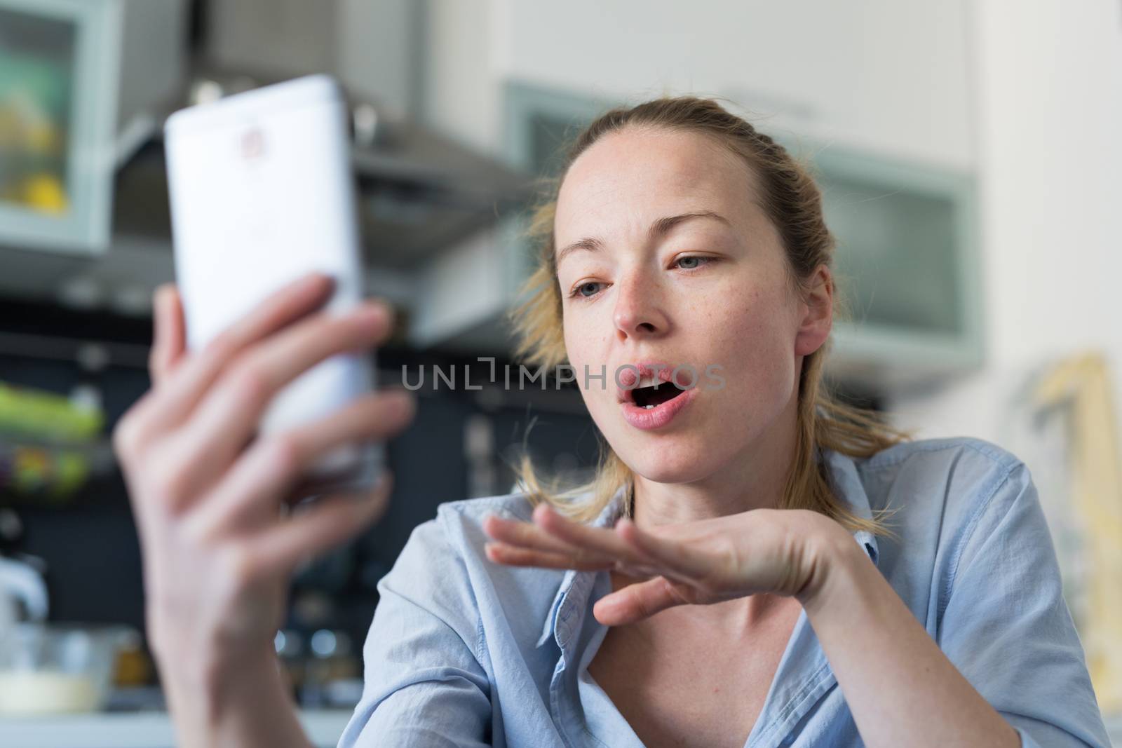 Young woman indoors at home kitchen using social media apps on phone for video chatting and stying connected with her loved ones. Stay at home, social distancing lifestyle.