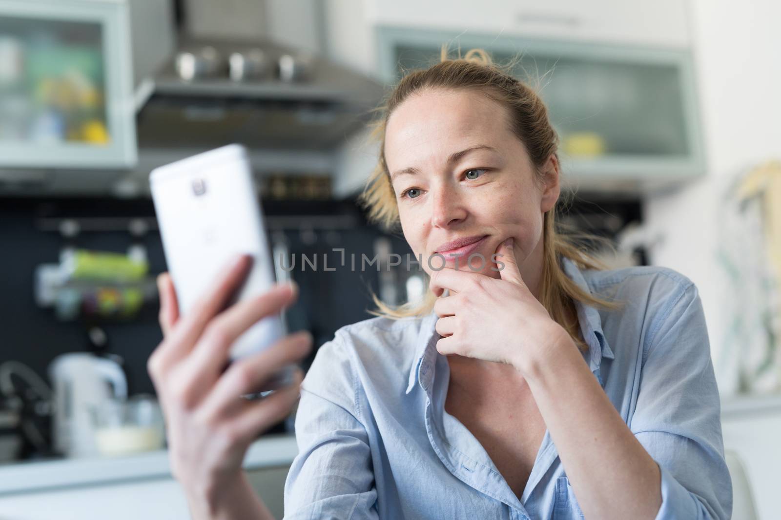 Young smiling cheerful woman indoors at home kitchen using social media apps on phone for video chatting and stying connected with her loved ones. Stay at home, social distancing lifestyle.