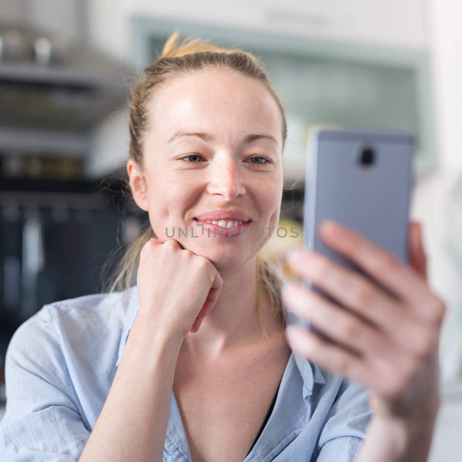 Young smiling cheerful pleased woman indoors at home kitchen using social media apps on mobile phone for chatting and stying connected with her loved ones. Stay at home, social distancing lifestyle. by kasto