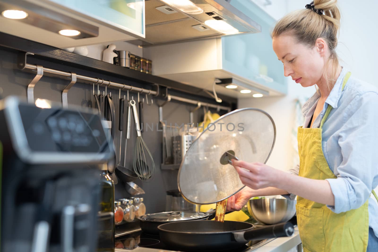 Stay at home housewife woman cooking in kitchen, stir frying dish in a saucepan, preparing food for family dinner. Blonde caucasian model in her 30is, real cooking not looking in camera.
