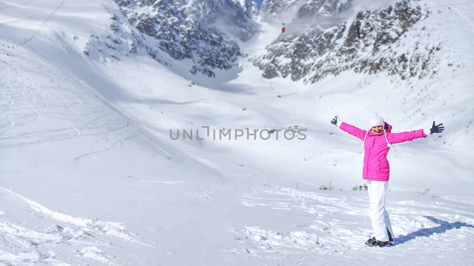 Happy young woman in pink ski jacket, gloves, hat and ski boots in front of snow covered mountain holding her hands spread out on a sunny day.