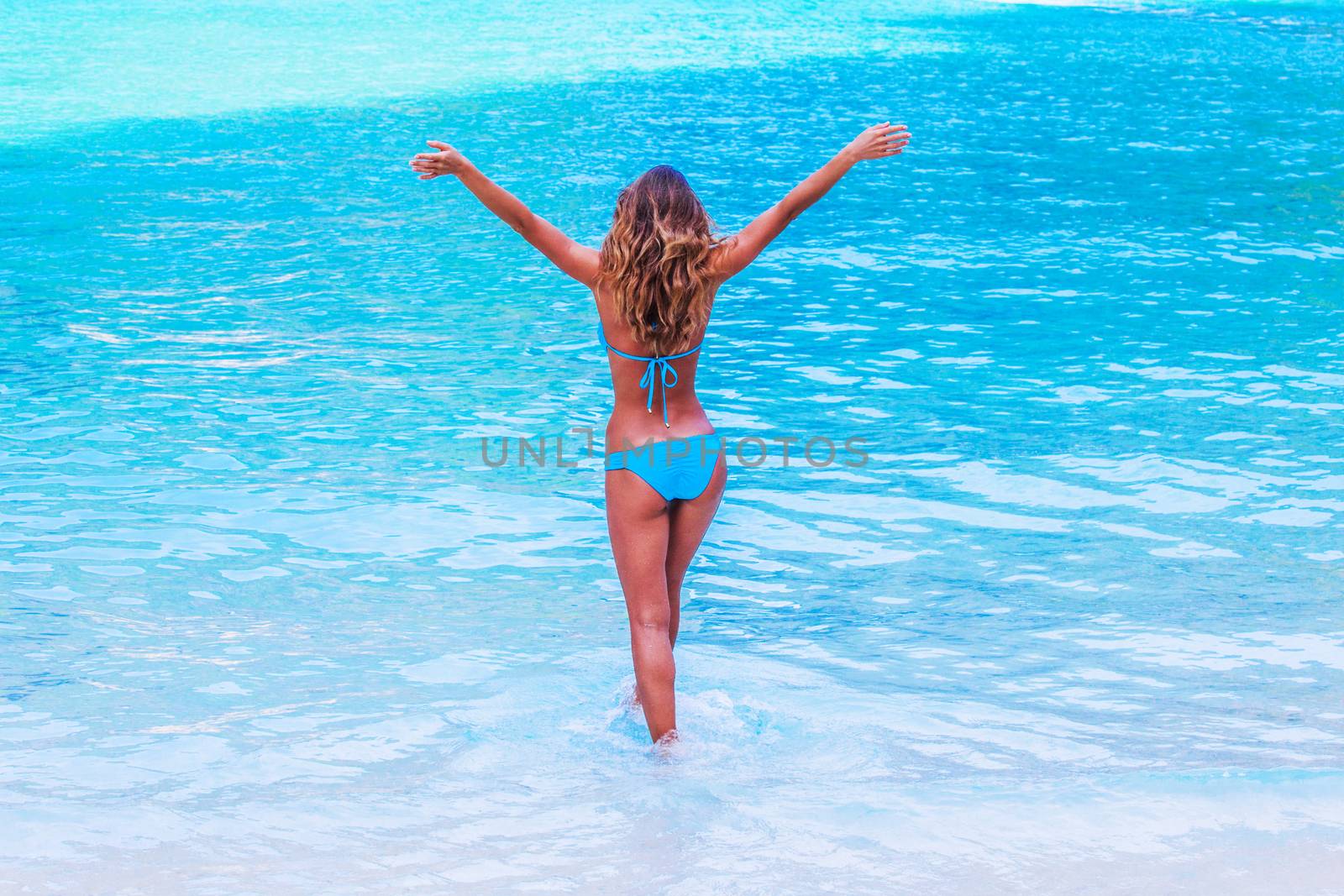 Beautiful woman in bikini walking to blue tropical sea with arms raised