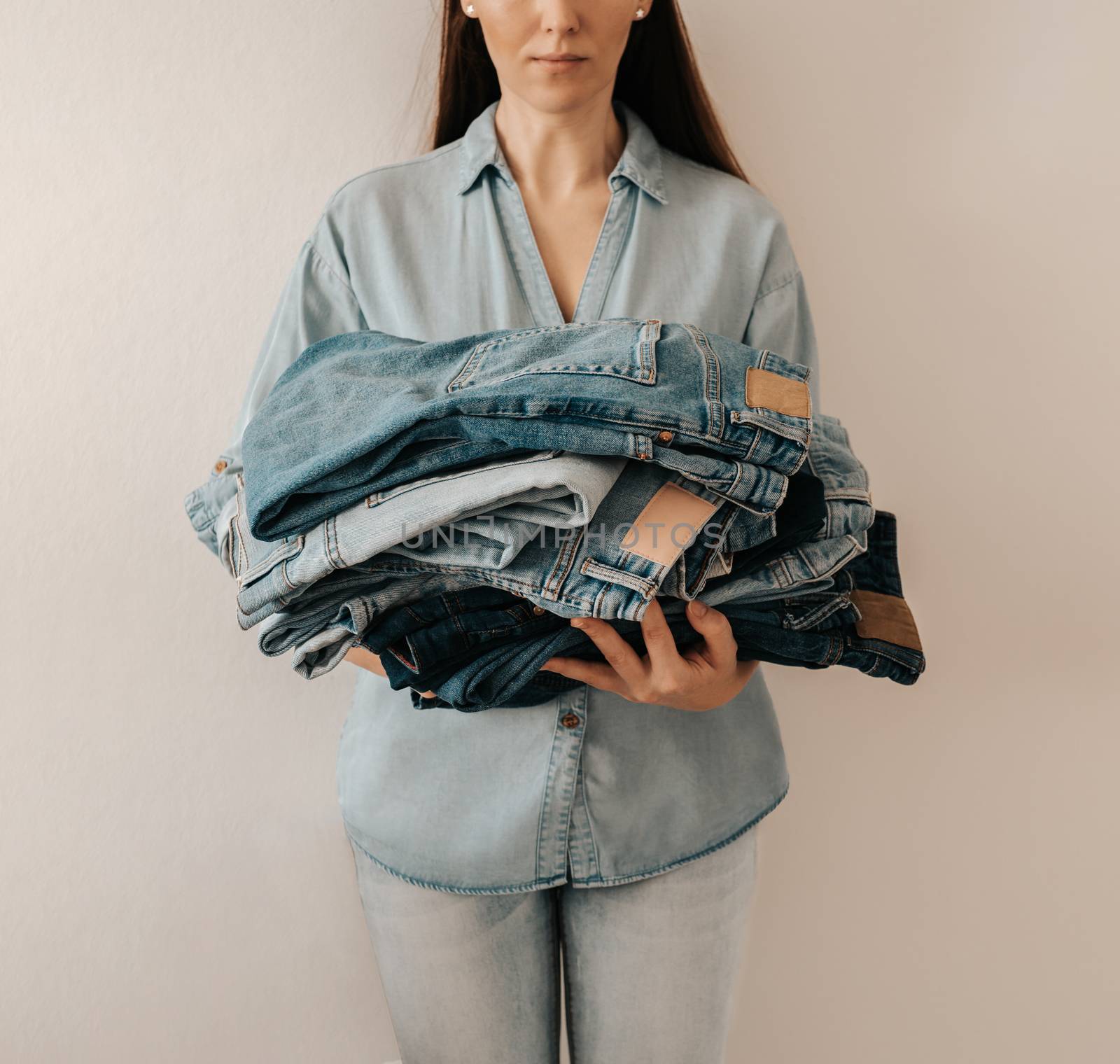 Unrecognizable woman holds stack of lot jeans pants on white background. Caucasian woman with long brown hair weared in blue shirt and jeans holds heap of denim pants with differents shades of blue