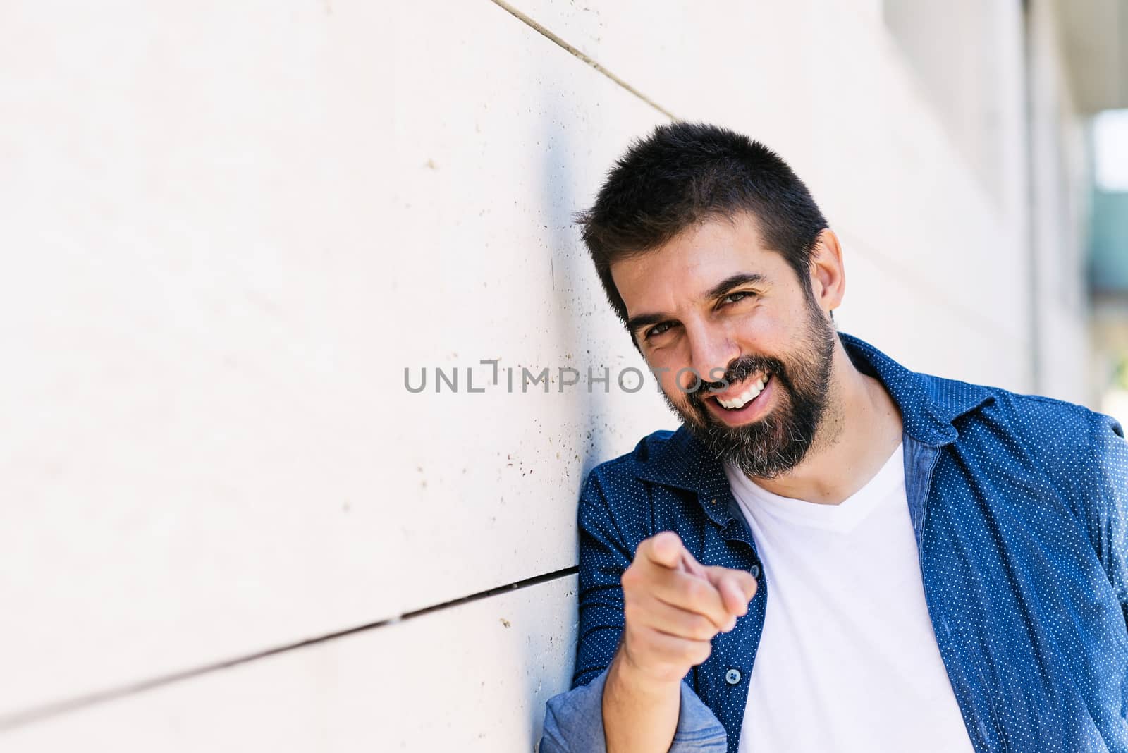 Bearded smiling male leaning on wall while pointing to you with finger