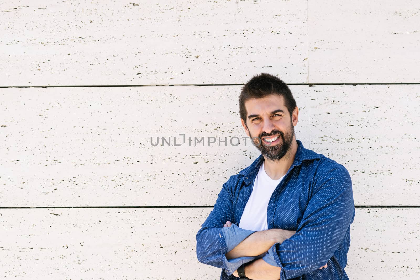 Bearded male leaning on wall with crossed arms while looking camera with a smile