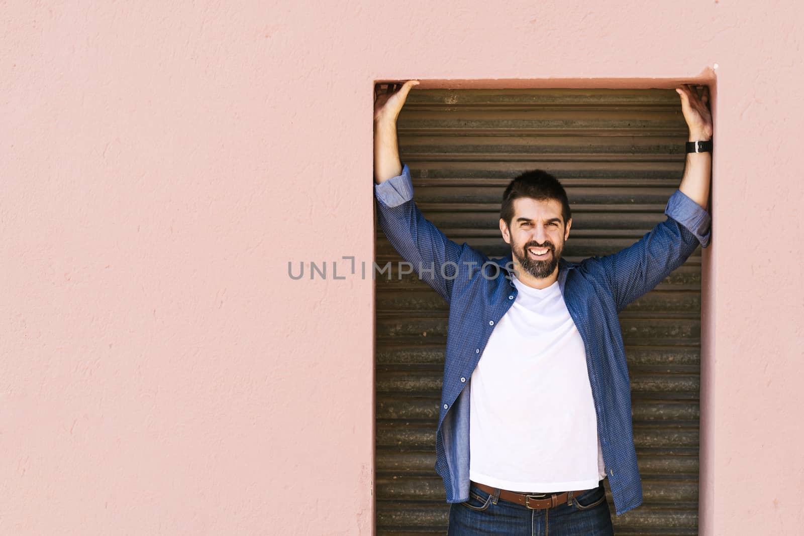 Bearded male standing against pink wall with arms raised while looking camera