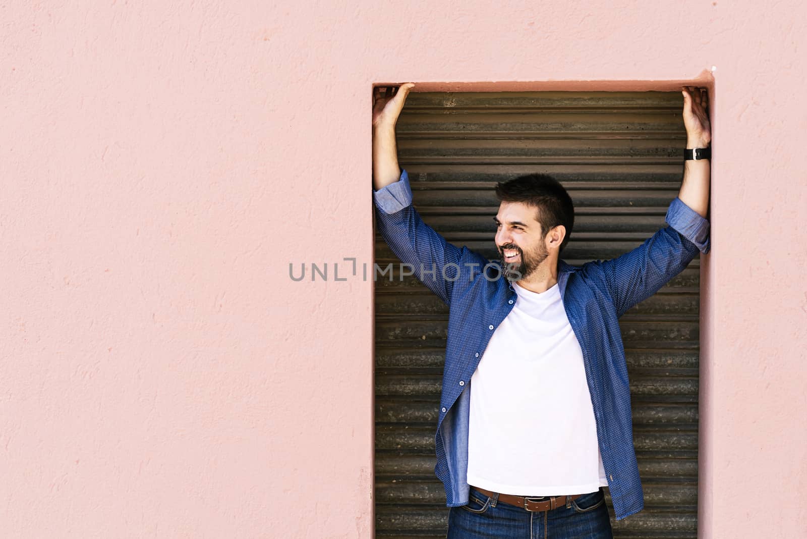 Bearded male standing against pink wall with arms raised while looking away
