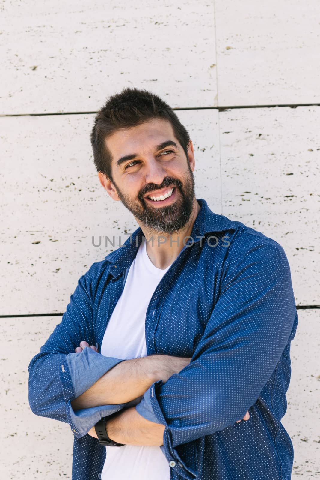 Bearded male leaning on wall with crossed arms while looking away with a smile