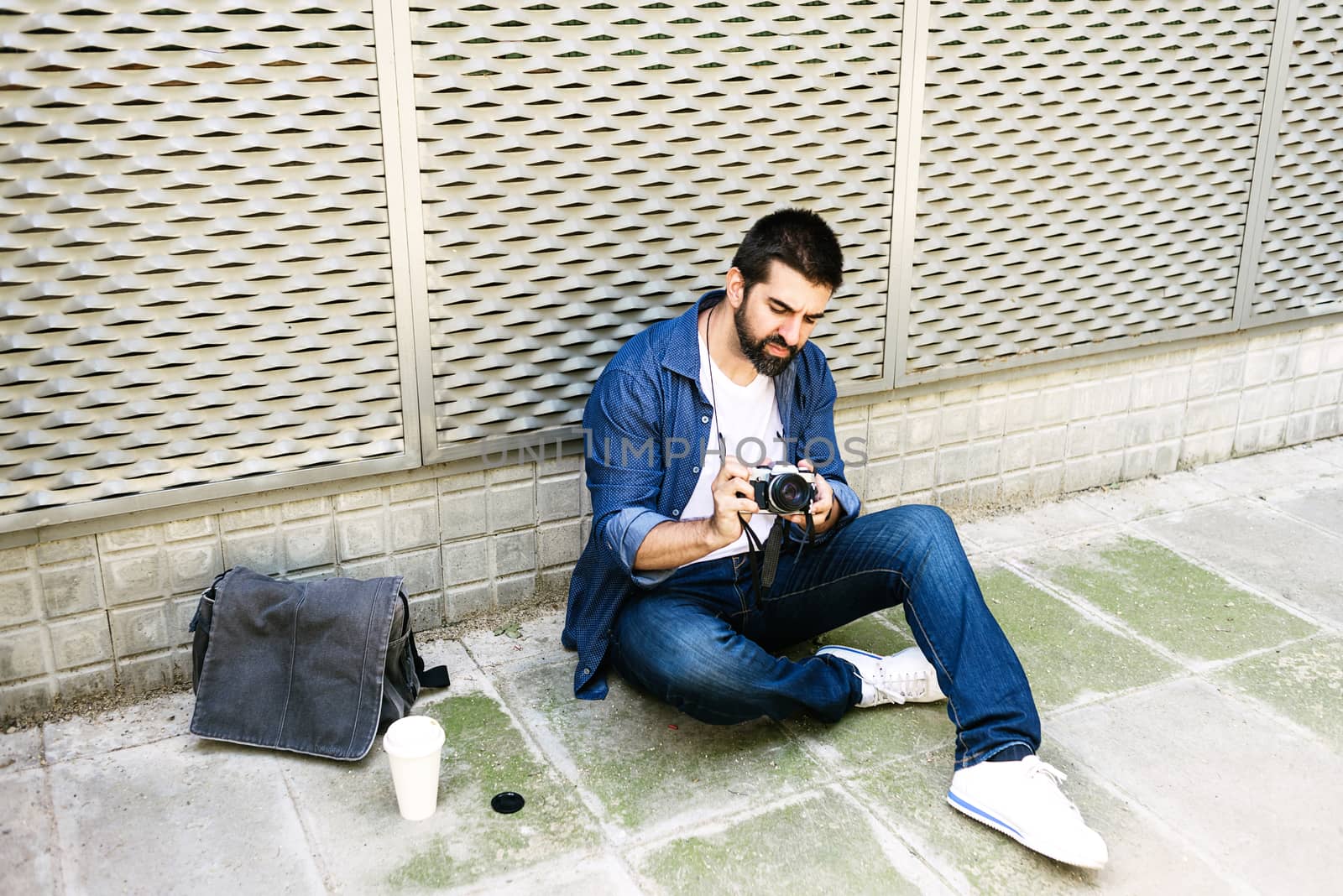 Cheerful bearded traveler man sitting on ground while using checking a camera