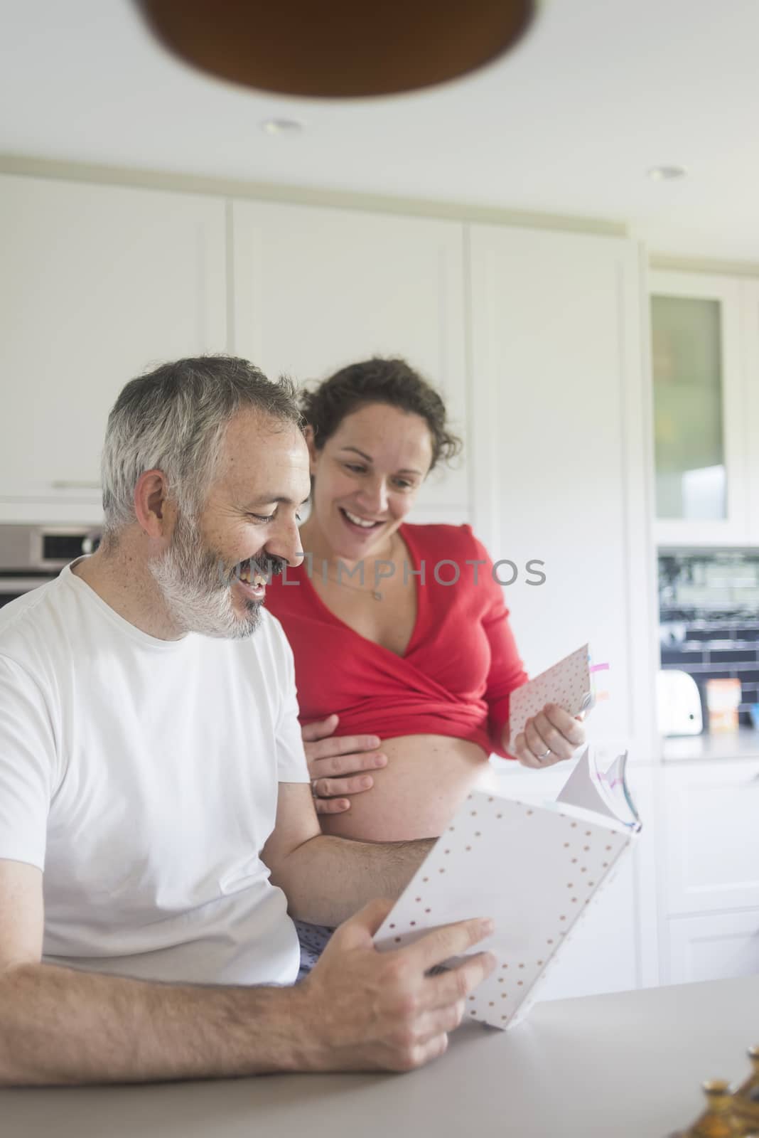 Happy pregnant woman with her husband sitting at home. Smiling couple consulting a book at the kitchen. by raferto1973