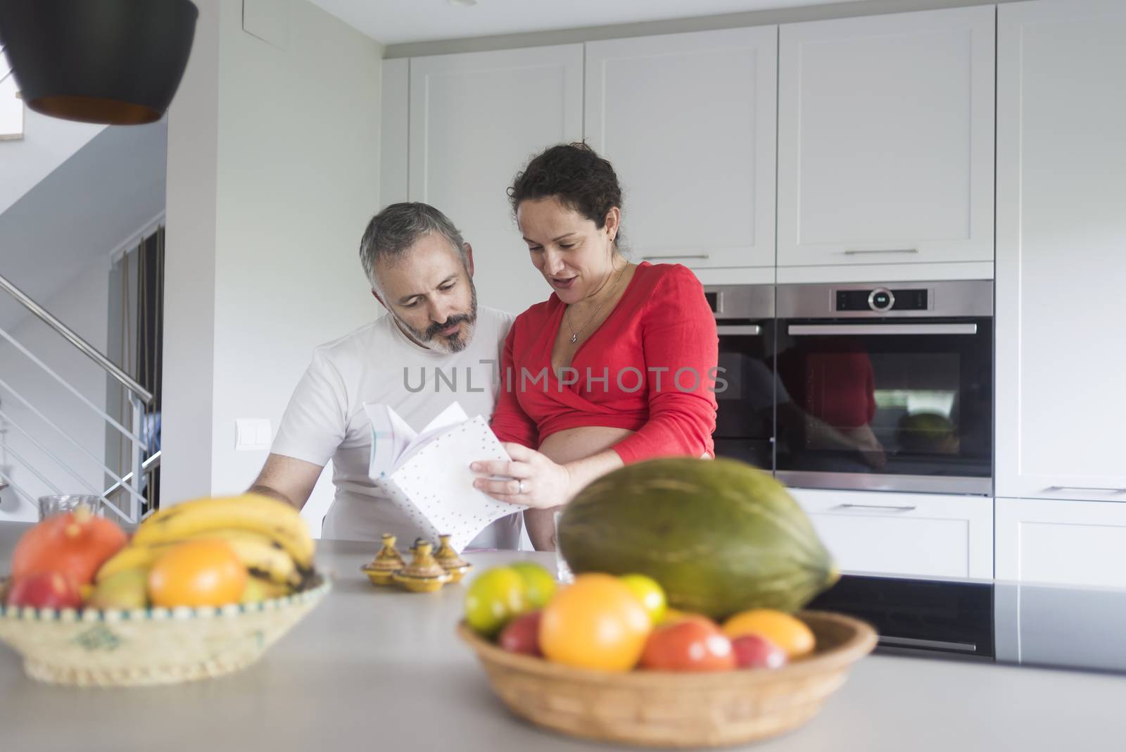 Happy pregnant woman with her husband sitting at home. Smiling couple consulting a book at the kitchen. by raferto1973