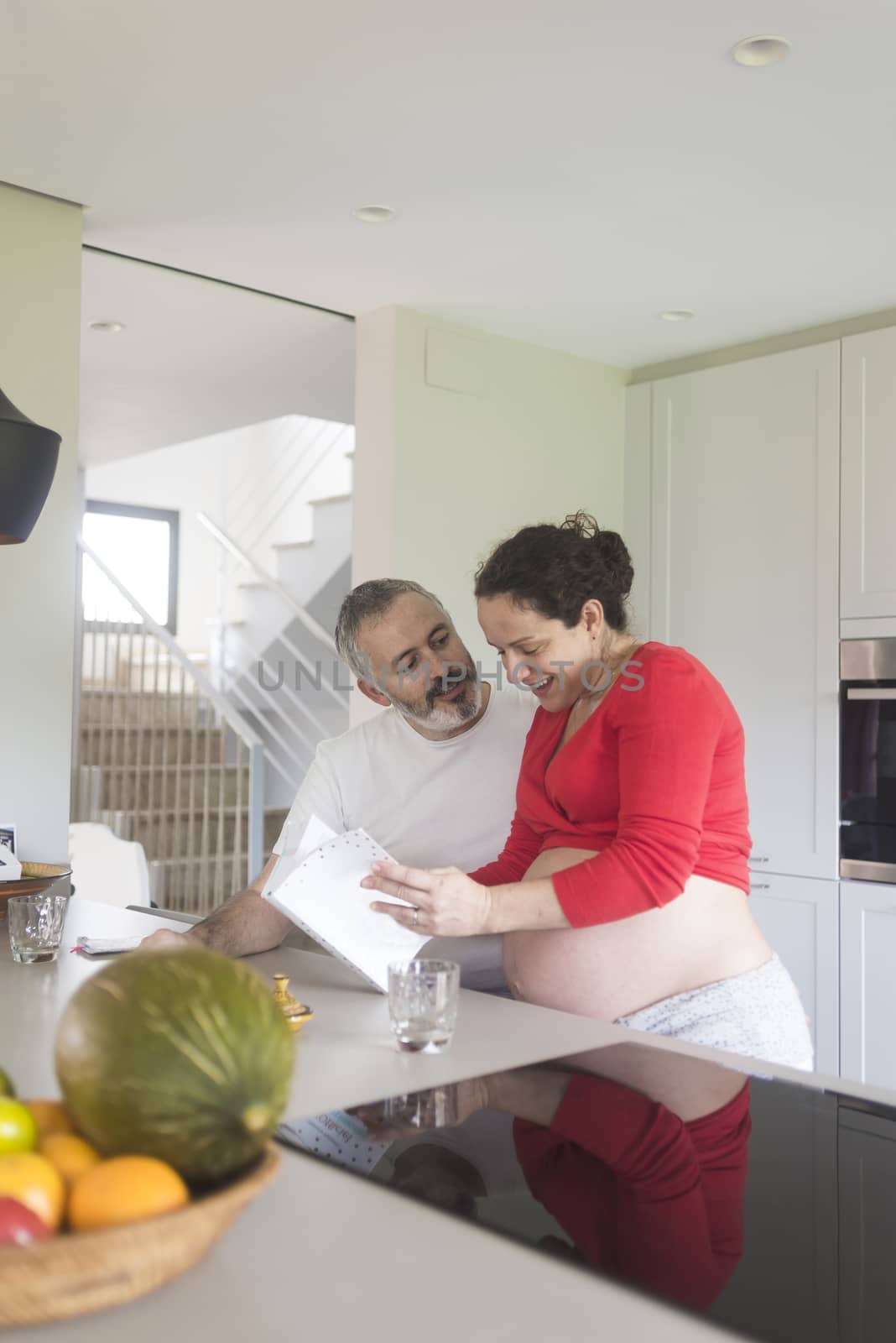 Happy pregnant woman with her husband sitting at home. Smiling couple consulting a book at the kitchen. by raferto1973