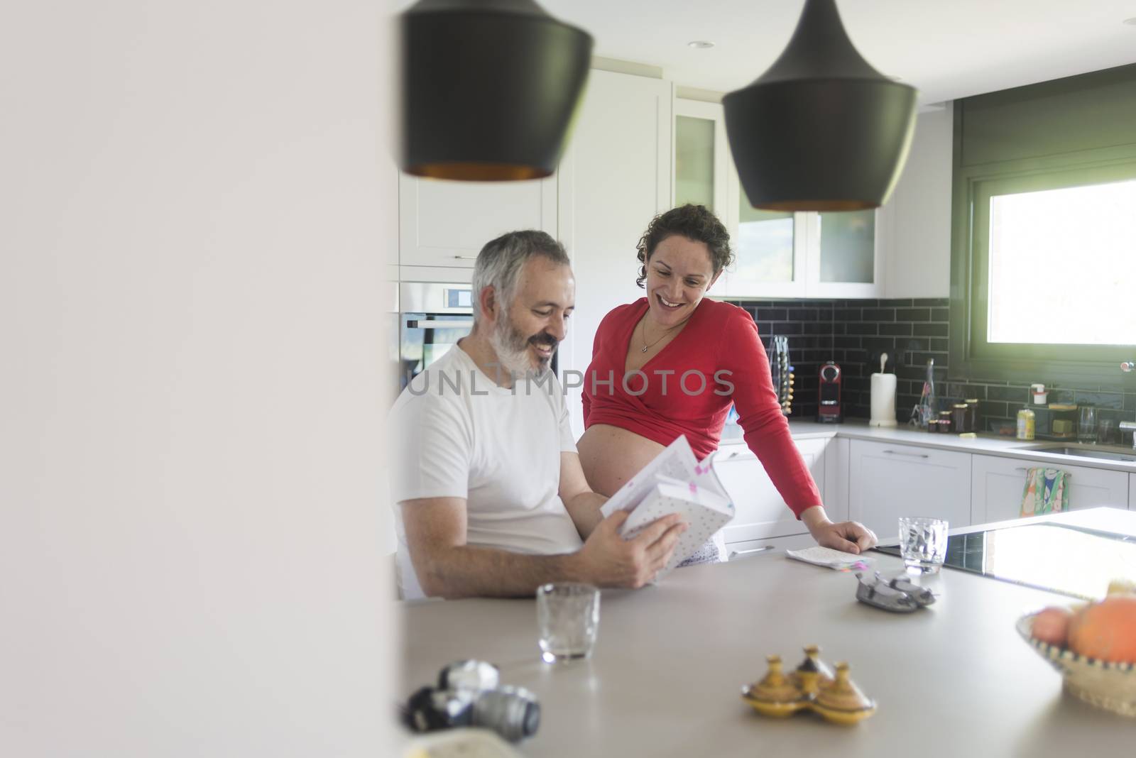 Happy pregnant woman with her husband sitting at home. Smiling couple consulting a book at the kitchen. by raferto1973