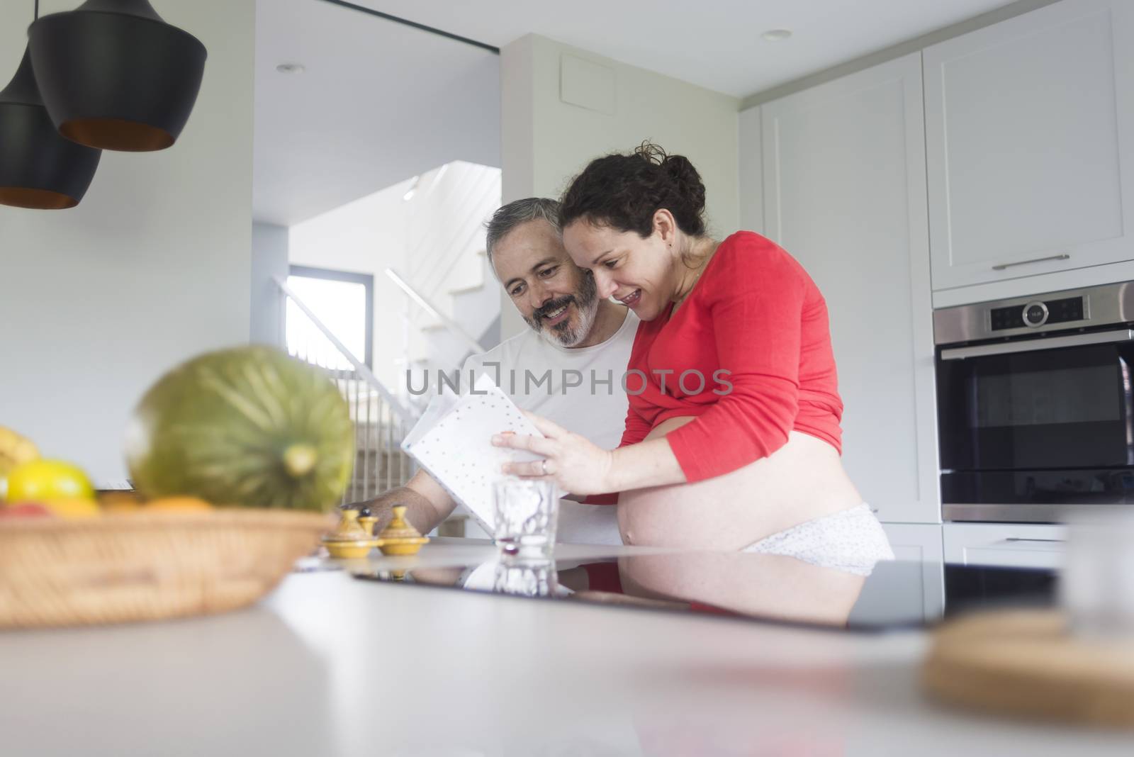 Happy pregnant woman with her husband sitting at home. Smiling couple consulting a book at the kitchen. by raferto1973