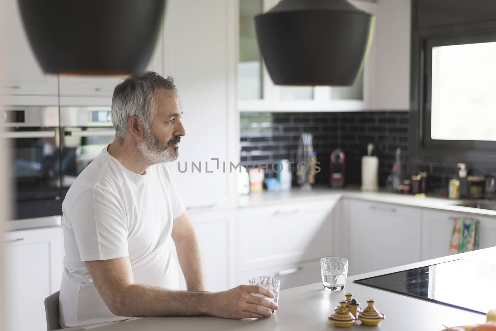 One bearded man sitting at the kitchen holding a glass of water while looking aside pensive