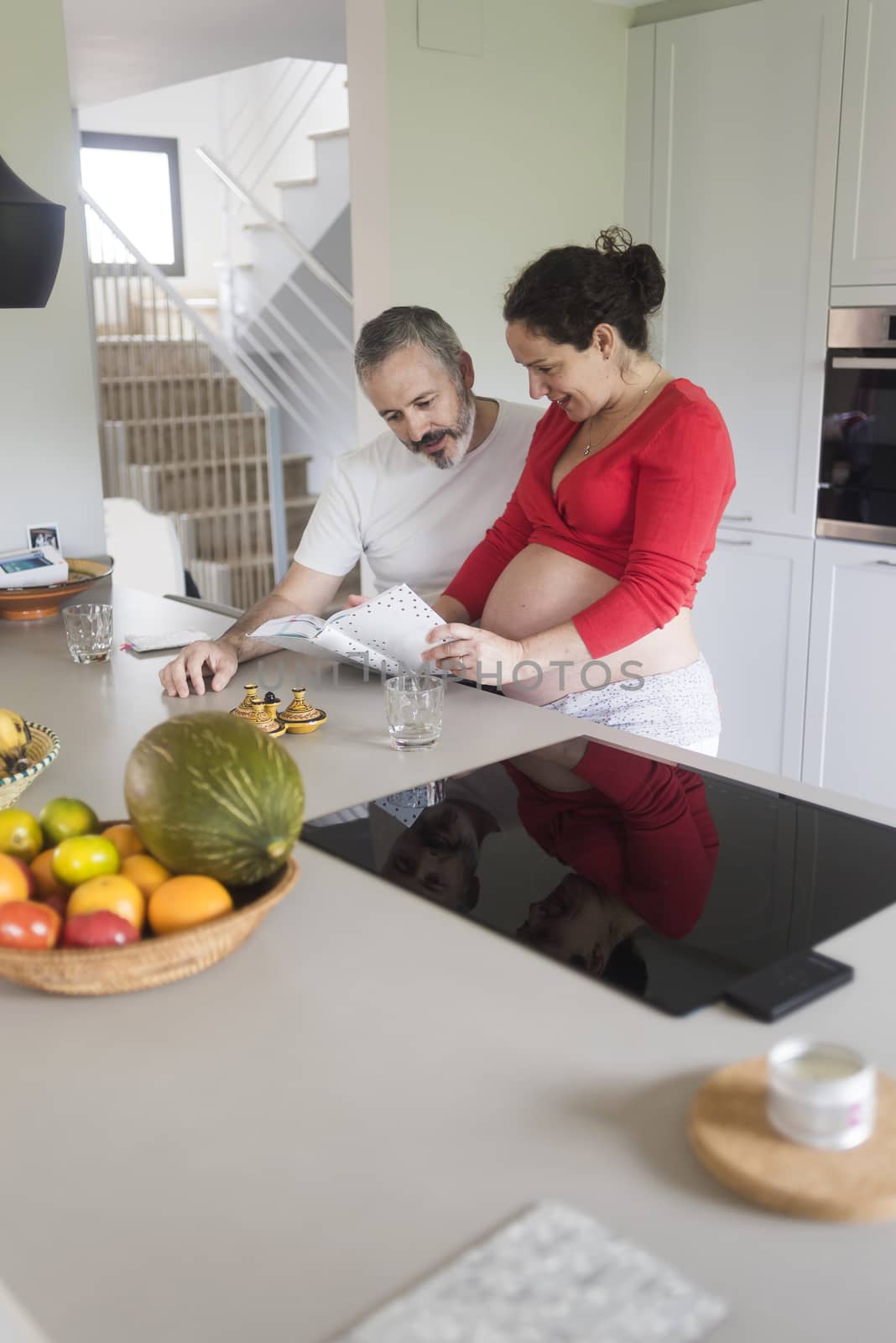 Happy pregnant woman with her husband sitting at home. Smiling couple consulting a book at the kitchen. by raferto1973