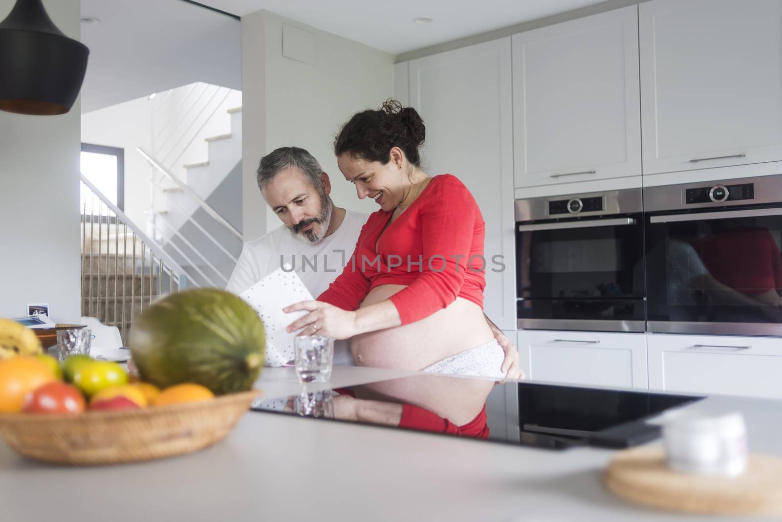 Happy pregnant woman with her husband sitting at home. Smiling couple consulting a book at the kitchen. by raferto1973