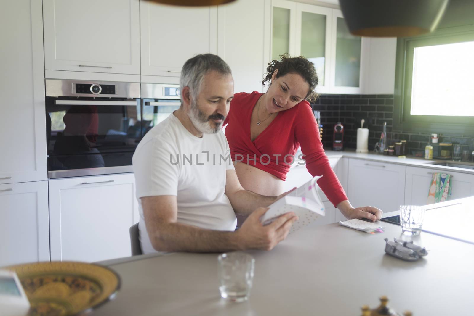 Happy pregnant woman with her husband sitting at home. Smiling couple consulting a book at the kitchen. by raferto1973