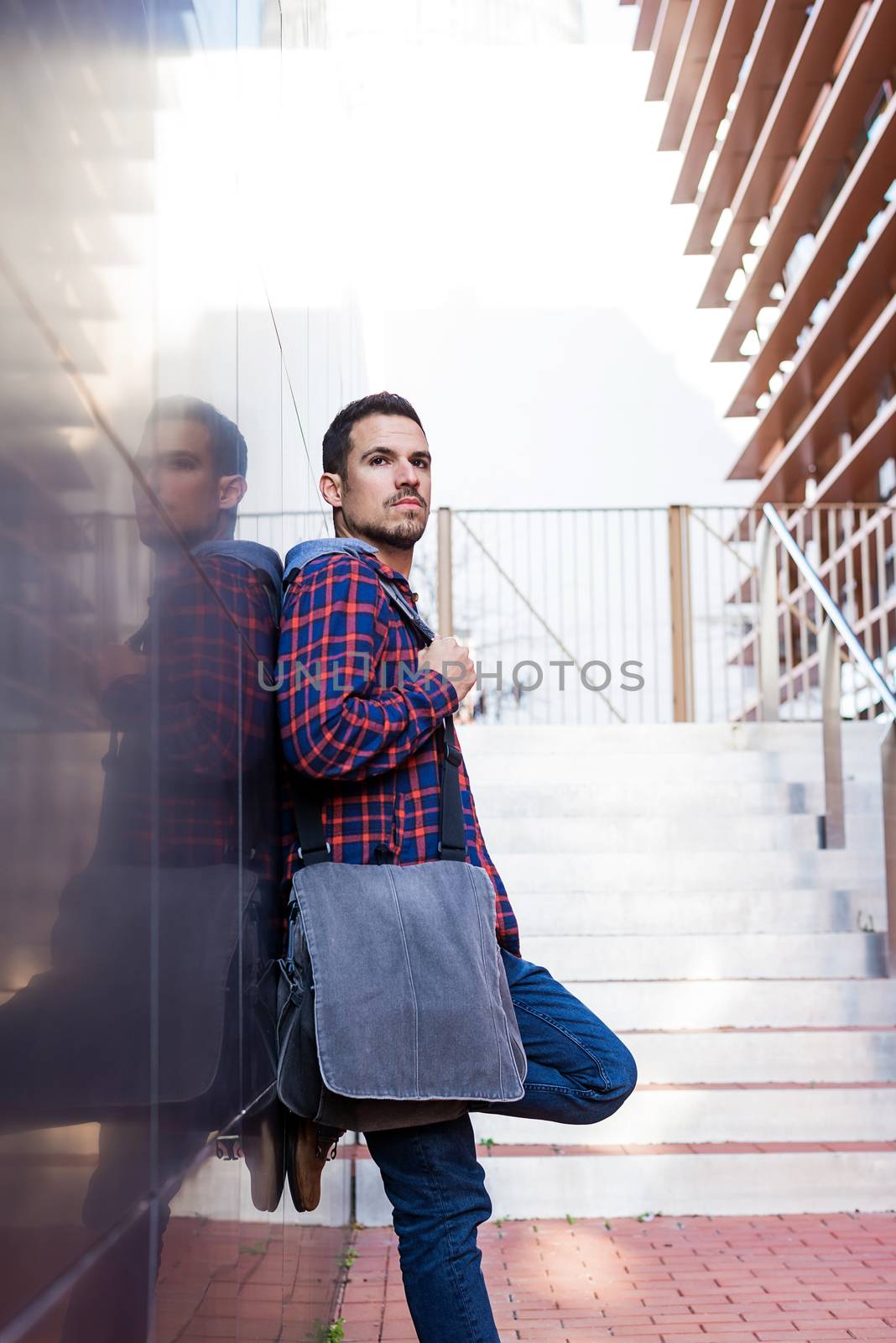 Young bearded guy leaning on a dark wall holding a shoulder bag while looking away