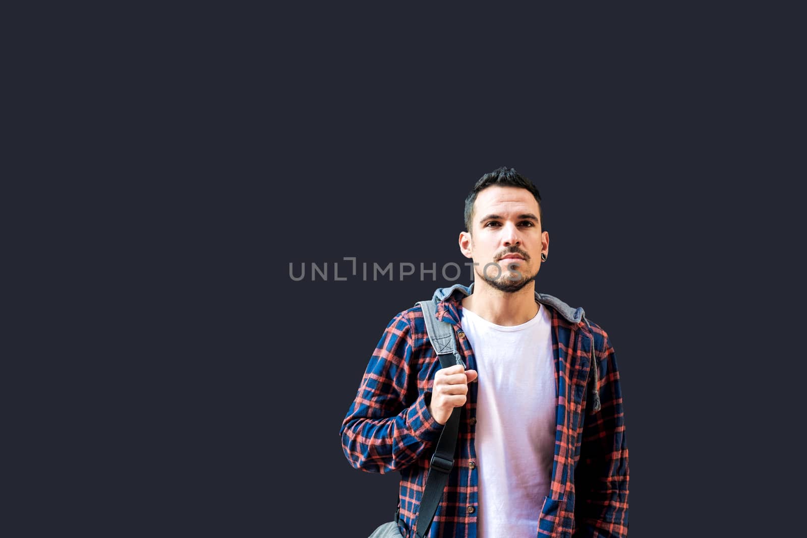 Young bearded guy leaning on a dark wall holding a shoulder bag while looking away