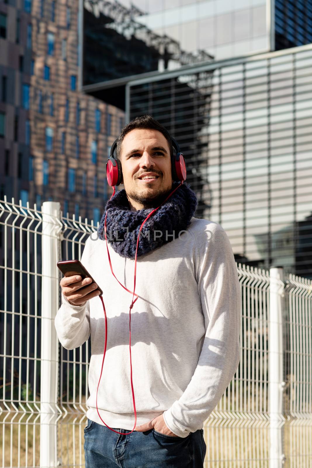 Young bearded man with headphones and holding smartphone while walking against skyscrapers in sunny day