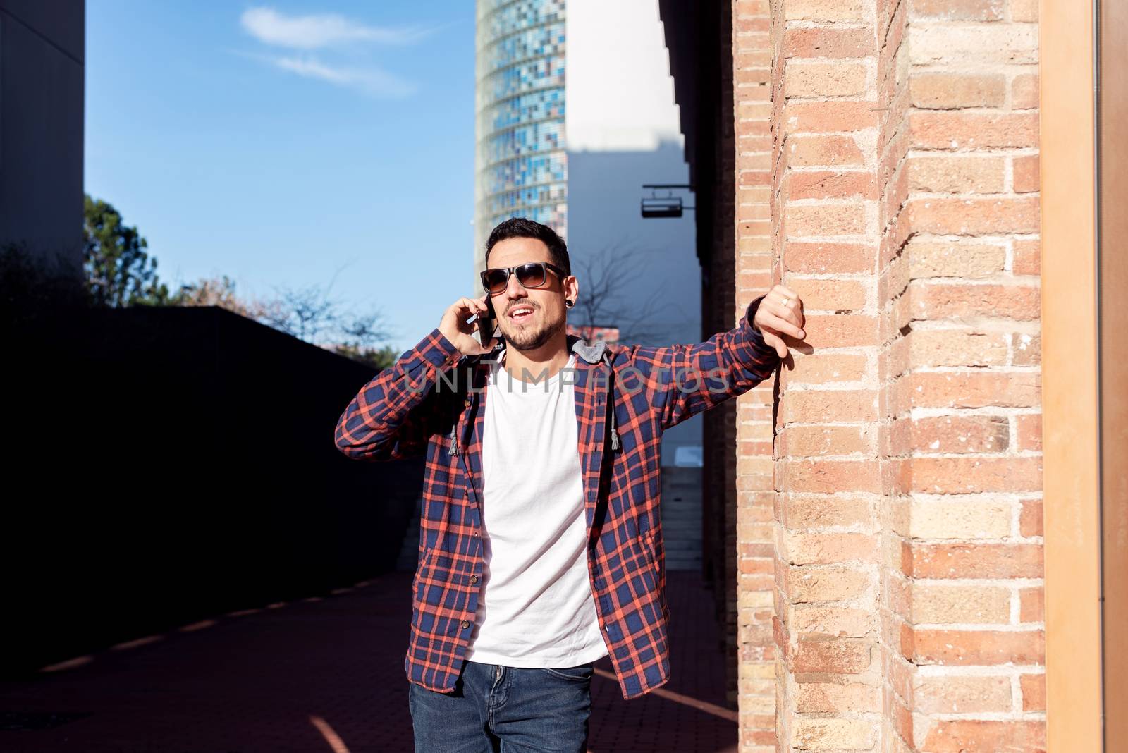 Young bearded man leaning on a bricked wall wearing sunglasses while using a smartphone outdoors. by raferto1973