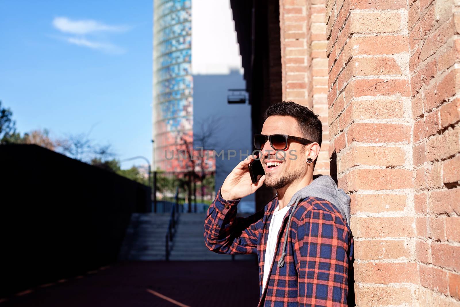 Young bearded male leaning on a bricked wall wearing sunglasses while using a smartphone outside.