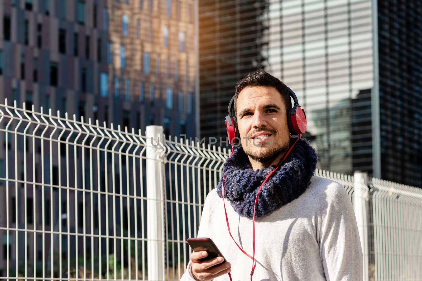 Young bearded man with headphones and holding smartphone while walking against skyscrapers in sunny day