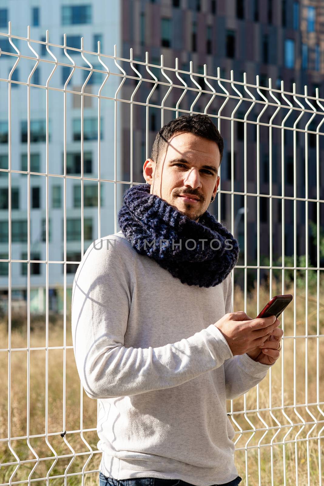 Young bearded man leaning on a metallic fence while using a smartphone