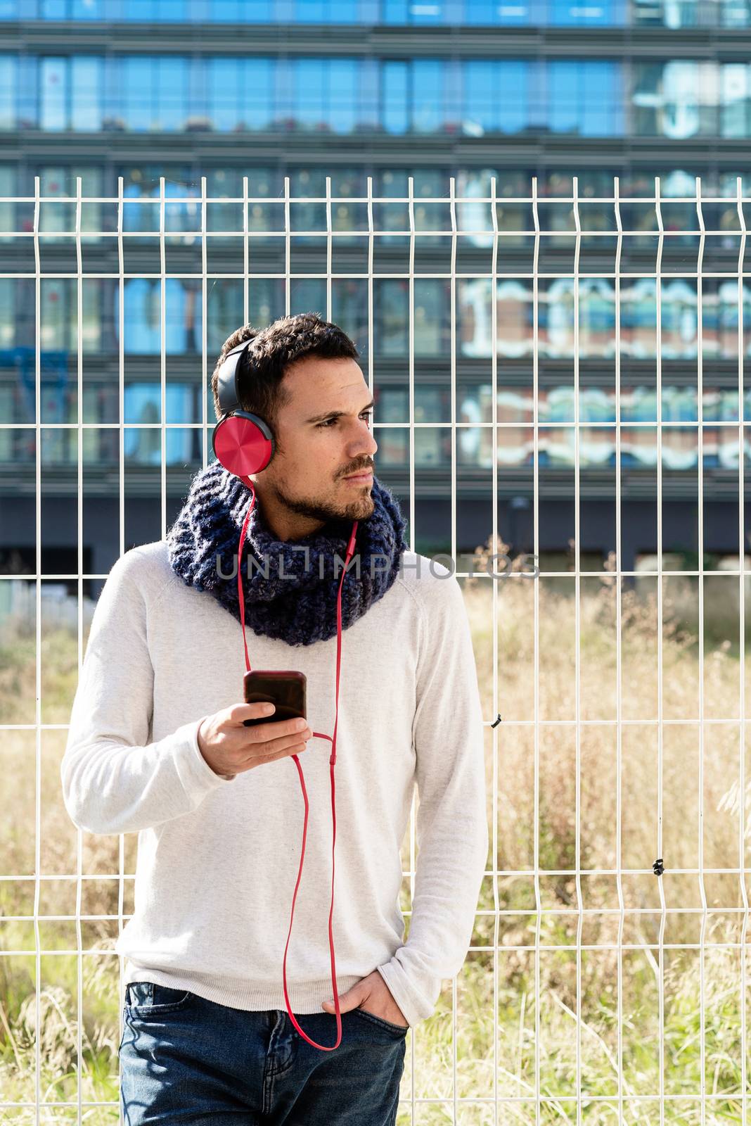 Young bearded male with headphones and holding smartphone while leaning on a metallic fence against skyscrapers in sunny day by raferto1973