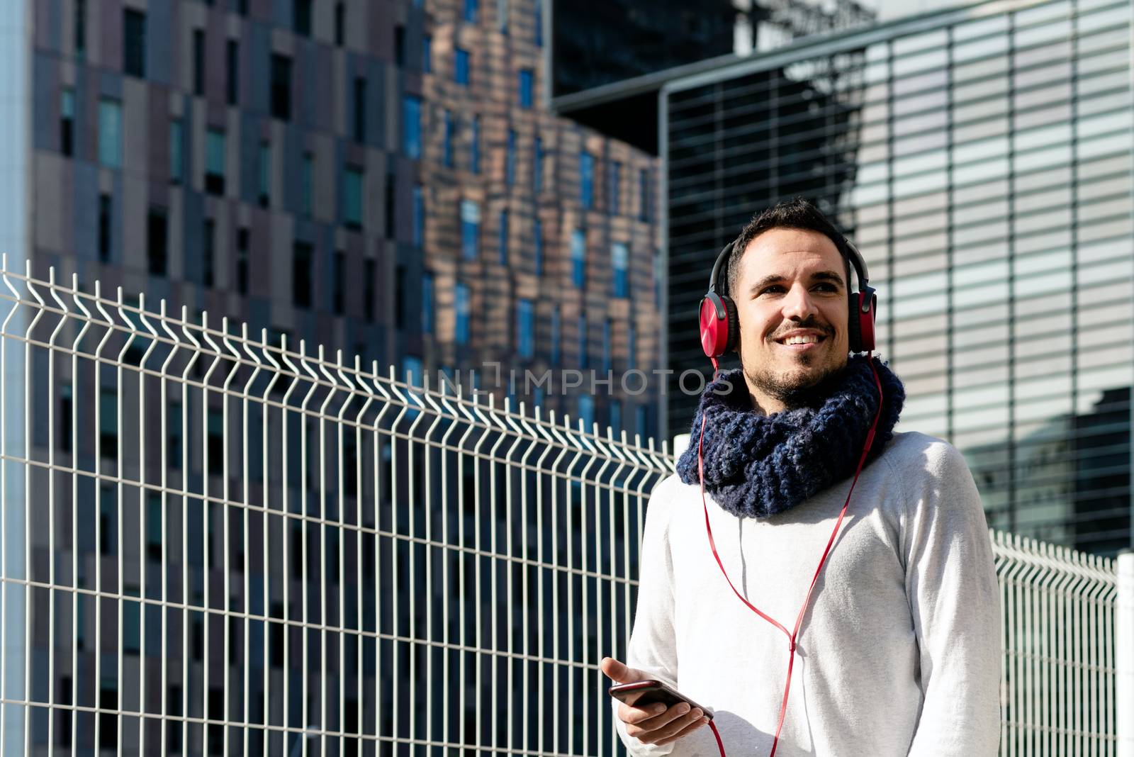 Young bearded man with headphones and holding smartphone while walking against skyscrapers in sunny day