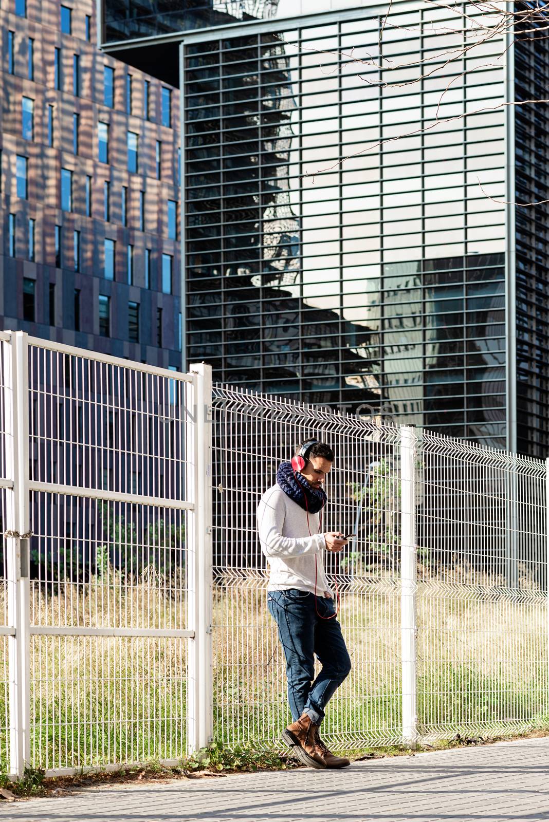 Young bearded male with headphones and holding smartphone while leaning on a metallic fence against skyscrapers in sunny day by raferto1973
