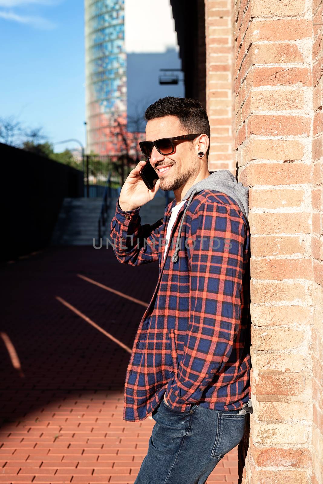 Young bearded male leaning on a bricked wall wearing sunglasses while using a smartphone outside.