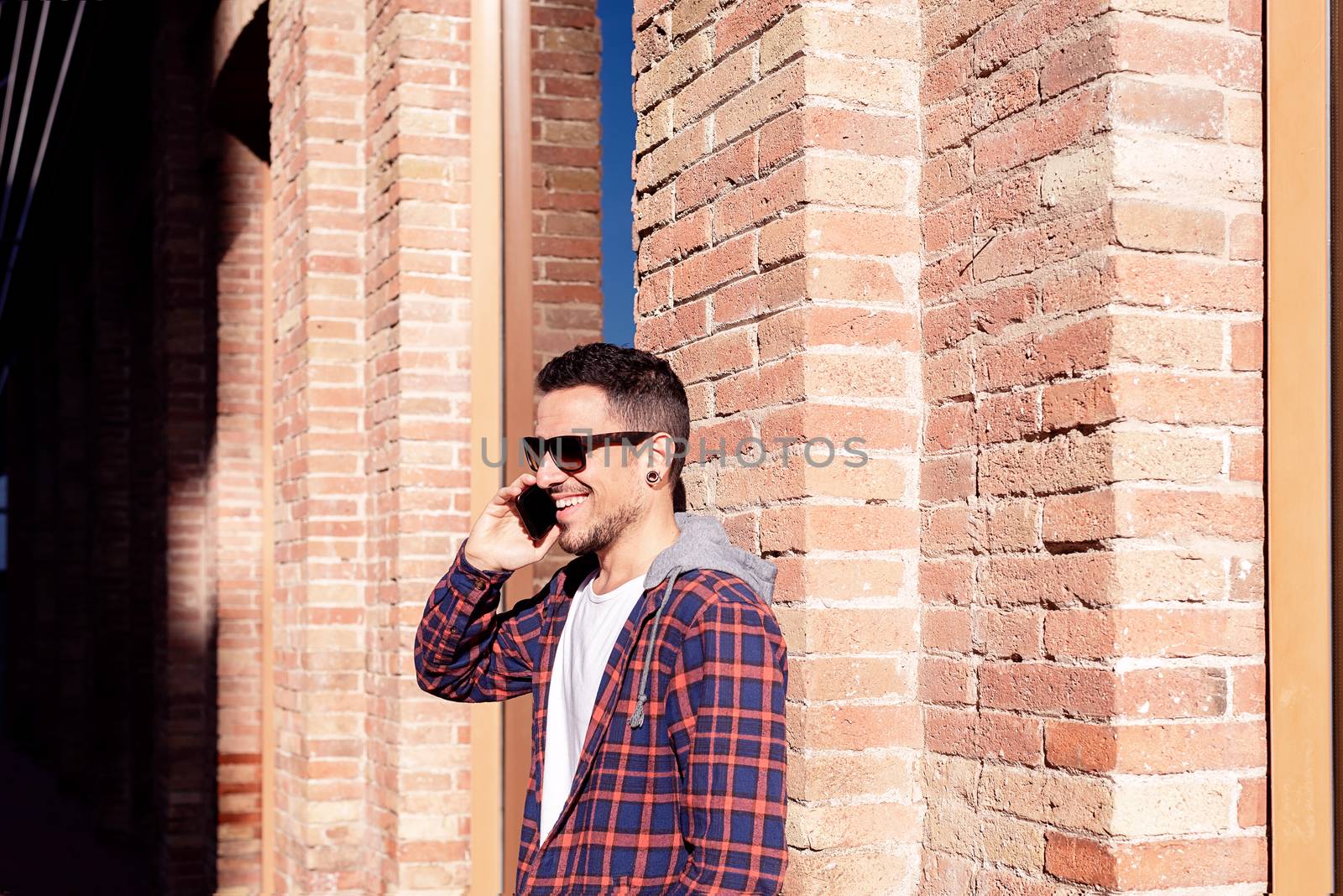 Young bearded man leaning on a bricked wall wearing sunglasses while using a smartphone outdoors. by raferto1973