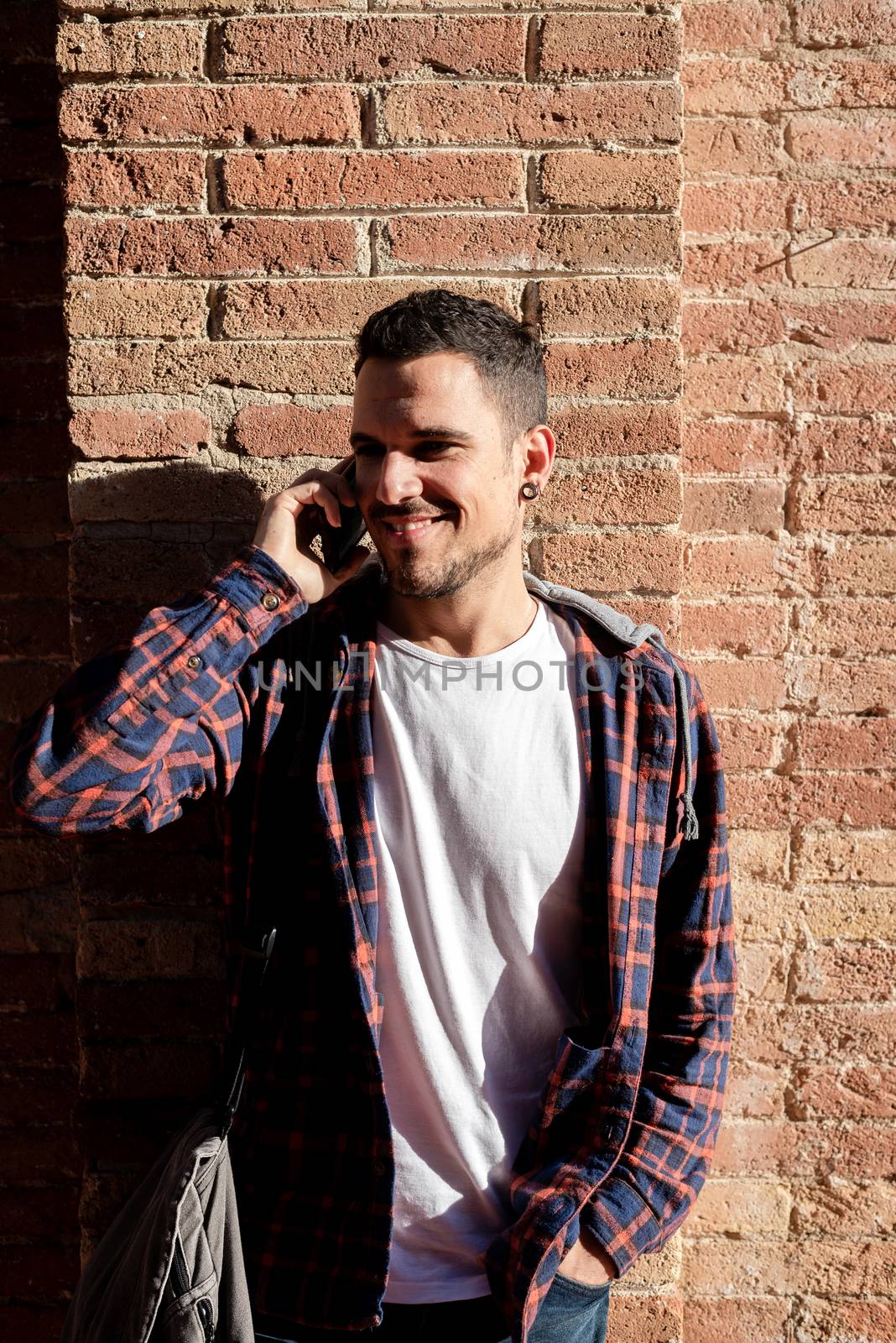 Young bearded man leaning on a bricked wall while using a smartphone outdoors. by raferto1973