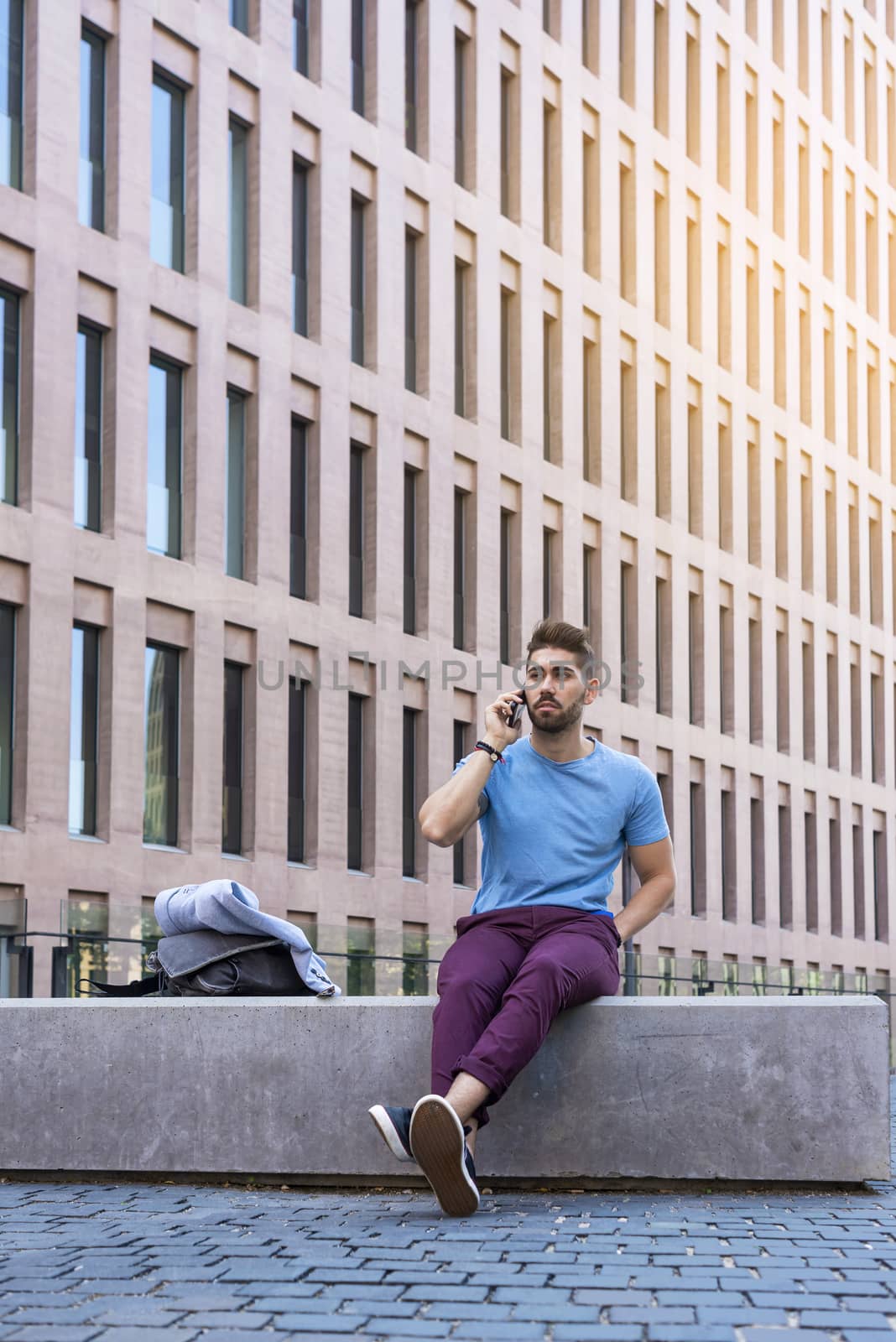 Portrait of cheerful modern businessman speaking by phone and smiling while sitting outdoors in a bench