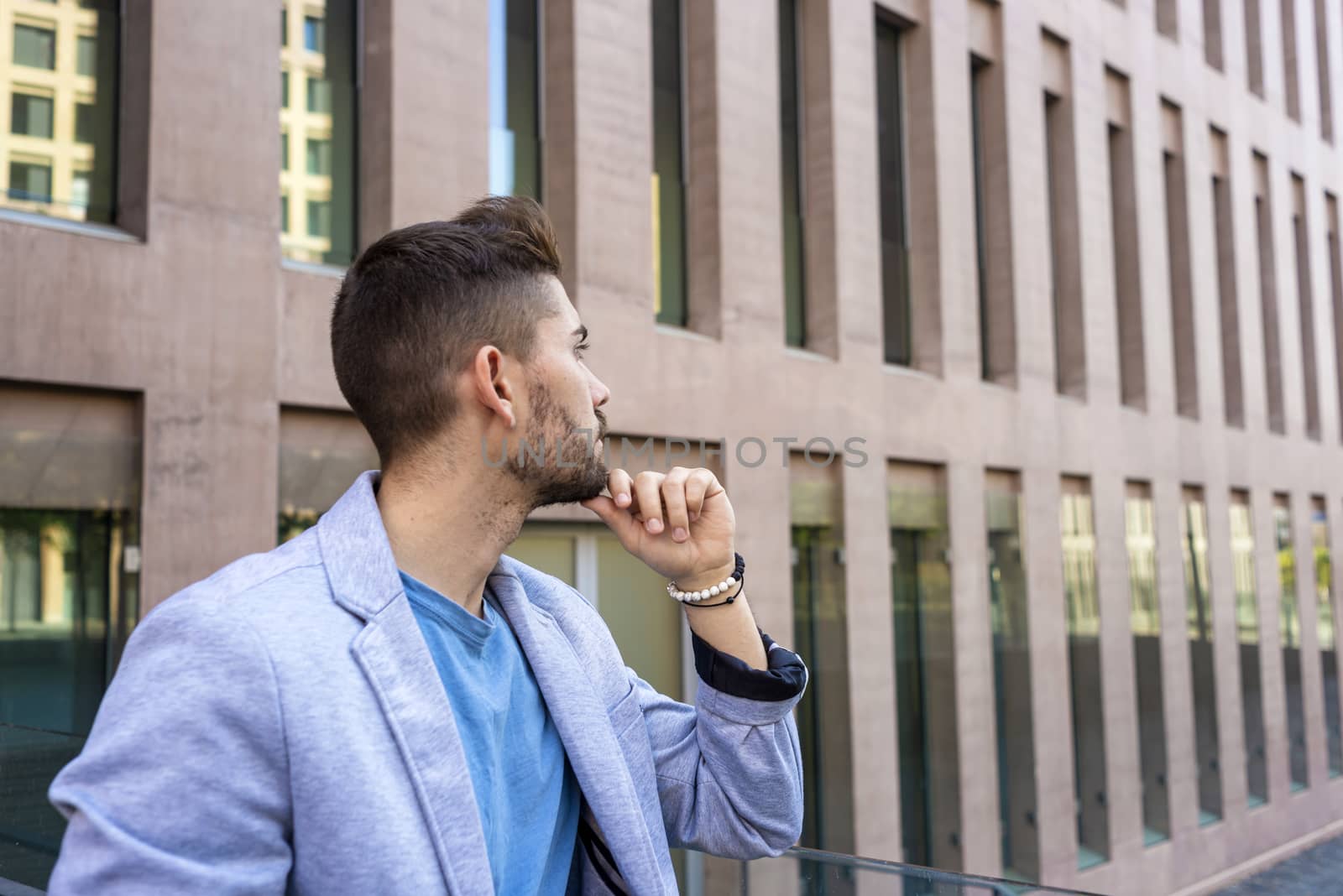 Bearded young man leaning on a fence with hand on chin while looking away