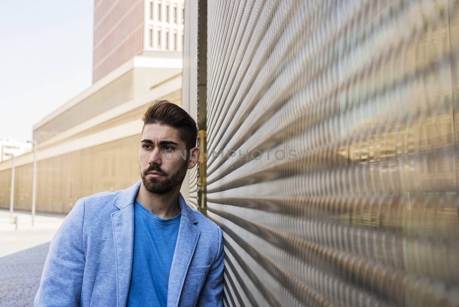 Young bearded man in urban background wearing casual clothes while leaning on a wall and looking aside