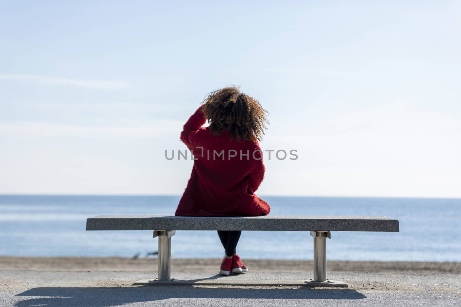 Rear view of a young curly woman wearing red denim jacket sitting on a bench while looking away to horizon over sea
